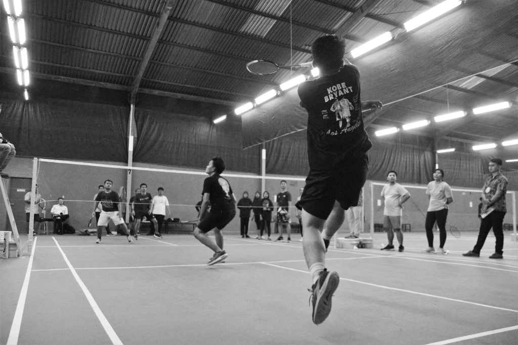Black and white image capturing intense indoor badminton match with players and spectators.
