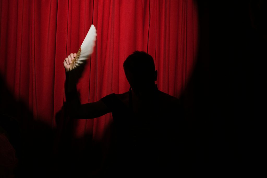 A silhouetted performer holds a fan in front of a vibrant red curtain, creating a dramatic stage scene.